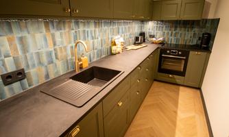 A modern kitchen with green cabinets and a stylish backsplash in blue and gray tones. The countertop is made of dark material and includes a sink as well as a cooking area.