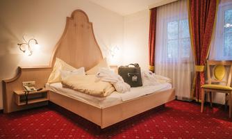 A cozy hotel room with a large bed and red carpet. Next to the bed is a chair and a bedside table with a phone.