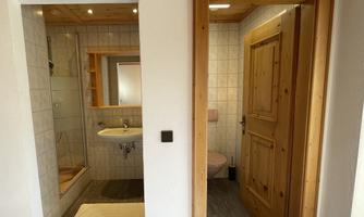 A modern bathroom with tiled floor and bright wood design. On the left side, there is a shower and a sink, on the right a door to another toilet.