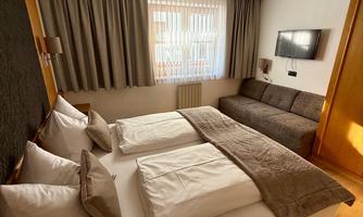 A modern hotel room with two beds and a cozy seating area. Bright windows provide natural light and a pleasant atmosphere.