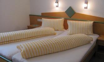 A simple bedroom with two single beds and striped pillows. The walls are light, with soft wall lamps and a small bedside table.