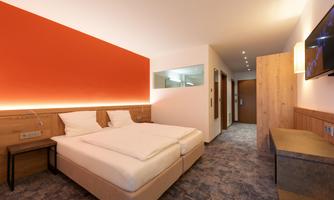 A modern hotel room with a double bed and an orange wall. The decor is elegant and filled with light.