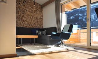 A modern living room with a wooden wall and a cozy sofa. Large windows provide a view of the mountains and plenty of daylight.