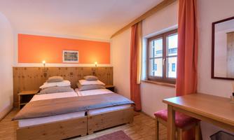 A cozy bedroom with two single beds and warm colors. The room has a large window and simple wooden furniture.