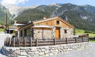 A typical alpine house made of stone and wood with a beautiful garden. In the background, high mountains and a clear blue sky can be seen.