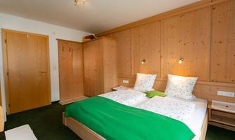 A cozy bedroom with two beds and a green bedspread. The walls are made of wood, and there is a dresser as well as a wardrobe.