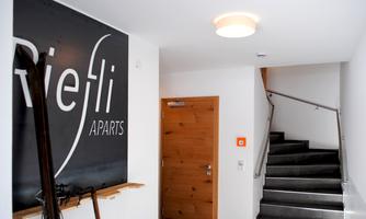 A bright hallway with a large wall board that says "Riefli Apparts." A wooden door and a staircase lead further up.
