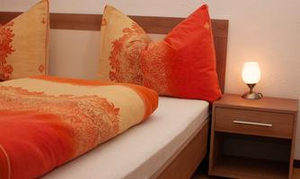 A cozy bedroom with a bed and bright orange pillows. Next to the bed is a wooden table with a small lamp.