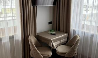A cozy area with a small table and two chairs. A television is mounted on the wall, while bright curtains let the light in.