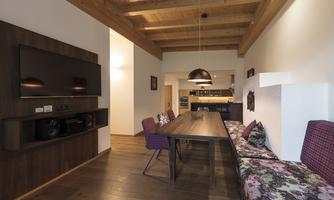 A modern living room with wooden ceilings and a large dining table. In the background, there is a cozy seating area with a floral pattern.