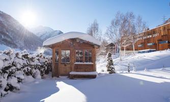 A cozy wooden house stands in a snow-covered landscape. In the background, majestic mountains rise under a radiant blue sky.