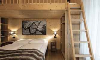 A cozy wooden bedroom with a double bed and a ladder to the loft. On the wall hangs a picture of two deer, and there is soft lighting from wall lamps.