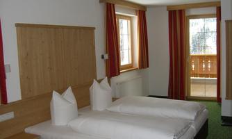 A cozy bedroom with two single beds and wooden furniture.  
Large windows with red curtains offer a view of a balcony.
