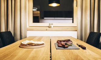 A modern wooden dining table with a plate of bread on the left side and a selection of meat on a black stone board on the right side. The room has a warm atmosphere with soft lighting.