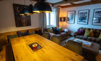 A cozy living space with a large wooden table and comfortable seating. The walls are decorated with artworks and photographs.