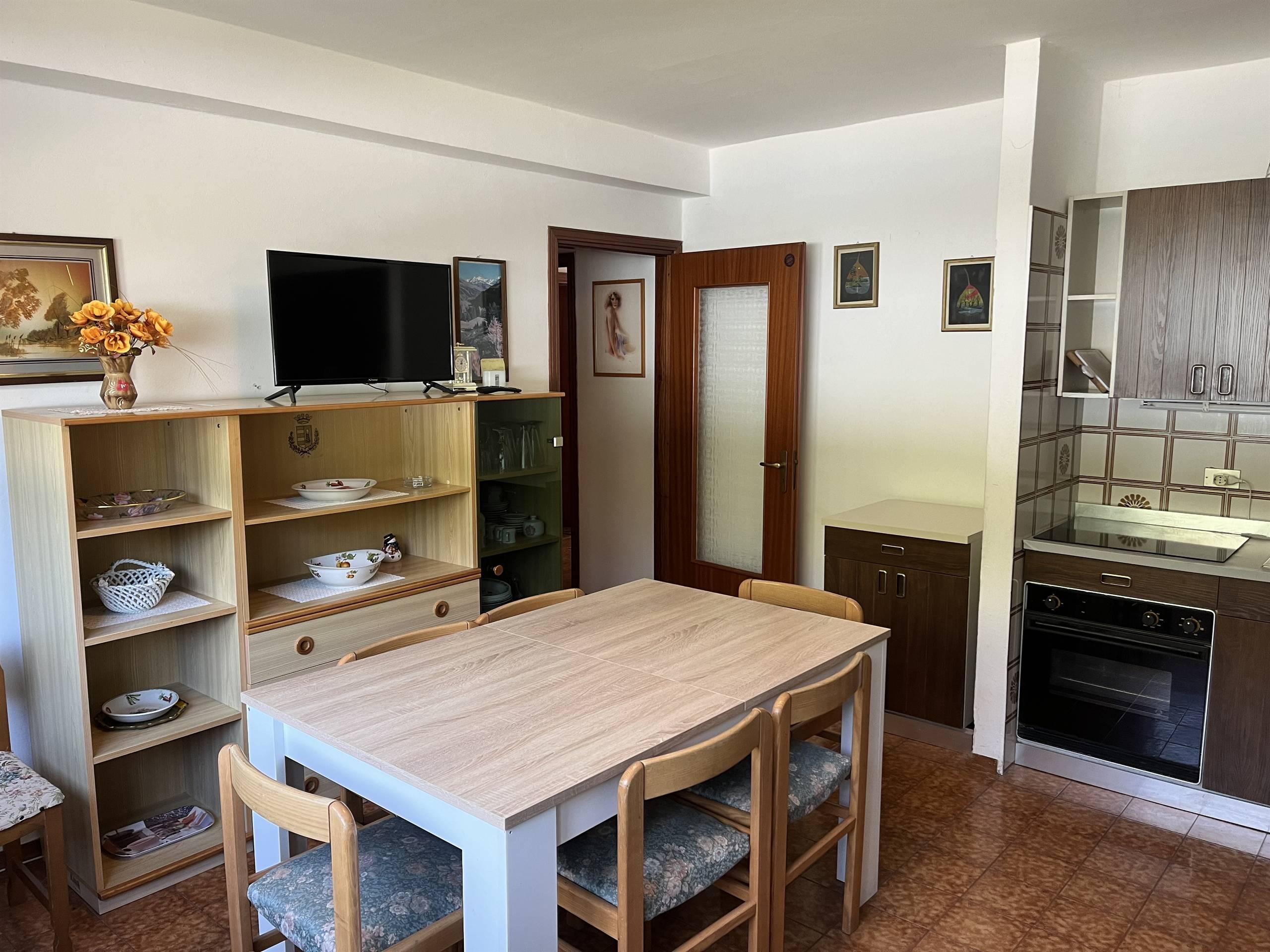 A cozy living room with a dining table and chairs. The room is furnished with a bookshelf, a television, and an open kitchen.