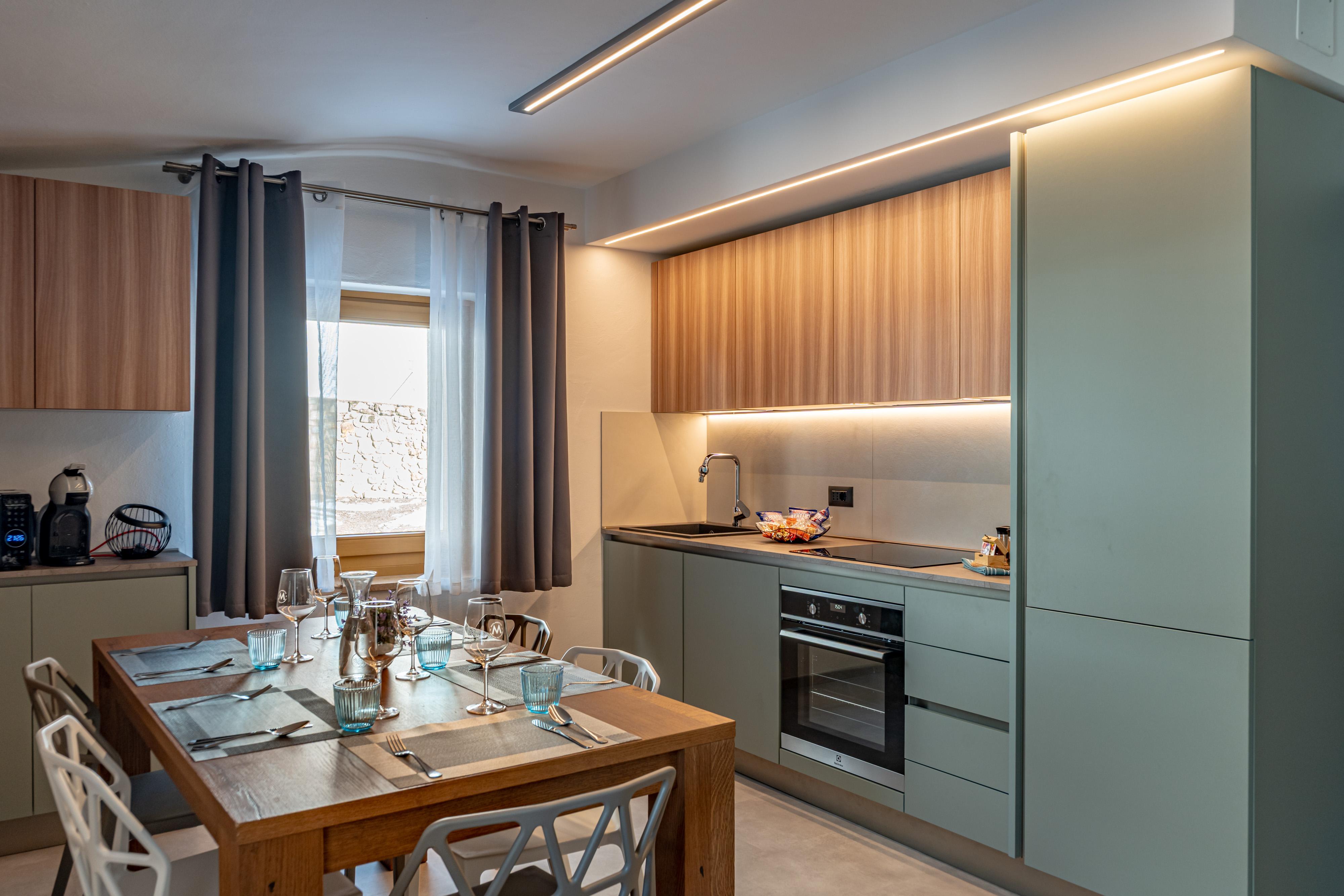 A modern kitchen with green cabinets and a large wooden table. The soft lighting creates a warm and inviting atmosphere.
