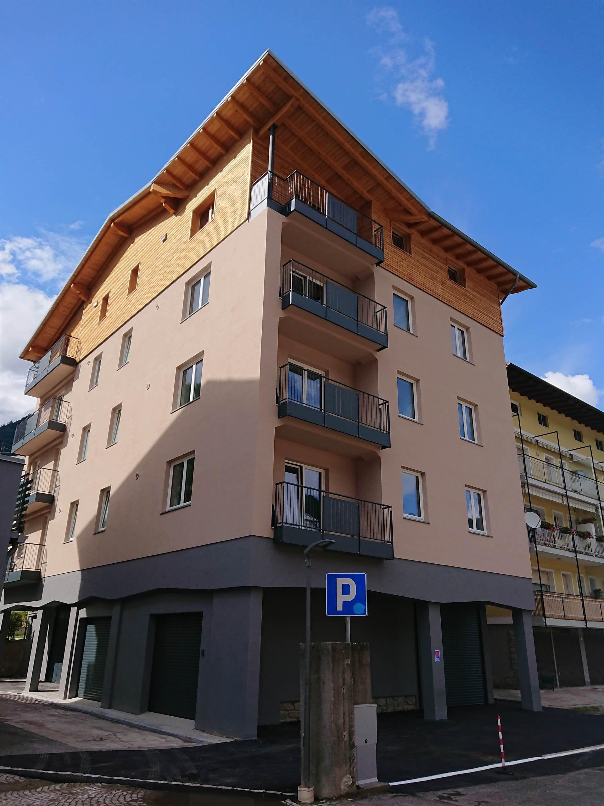 A modern three-story building with balconies and a light-colored facade. In front, there is a designated parking area.