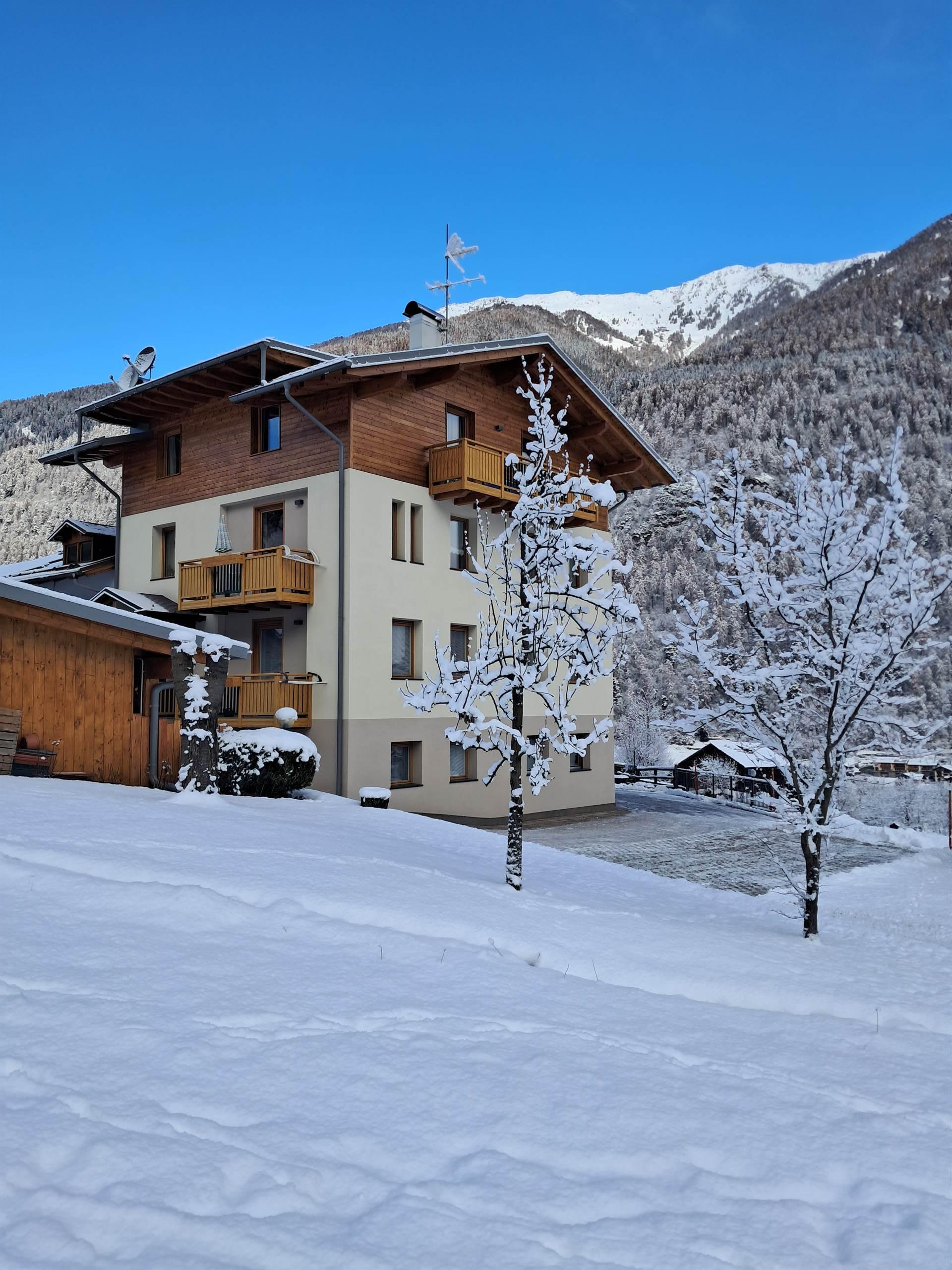 Una casa circondata da neve fresca, con alberi innevati e montagne sullo sfondo. Il cielo è sereno e blu.