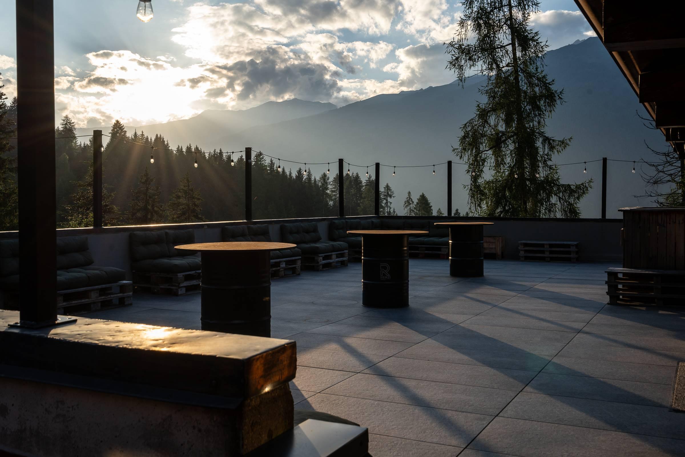 An elegant terrace with wooden tables, immersed in nature. In the background, mountains and a clear sky at sunset can be seen.
