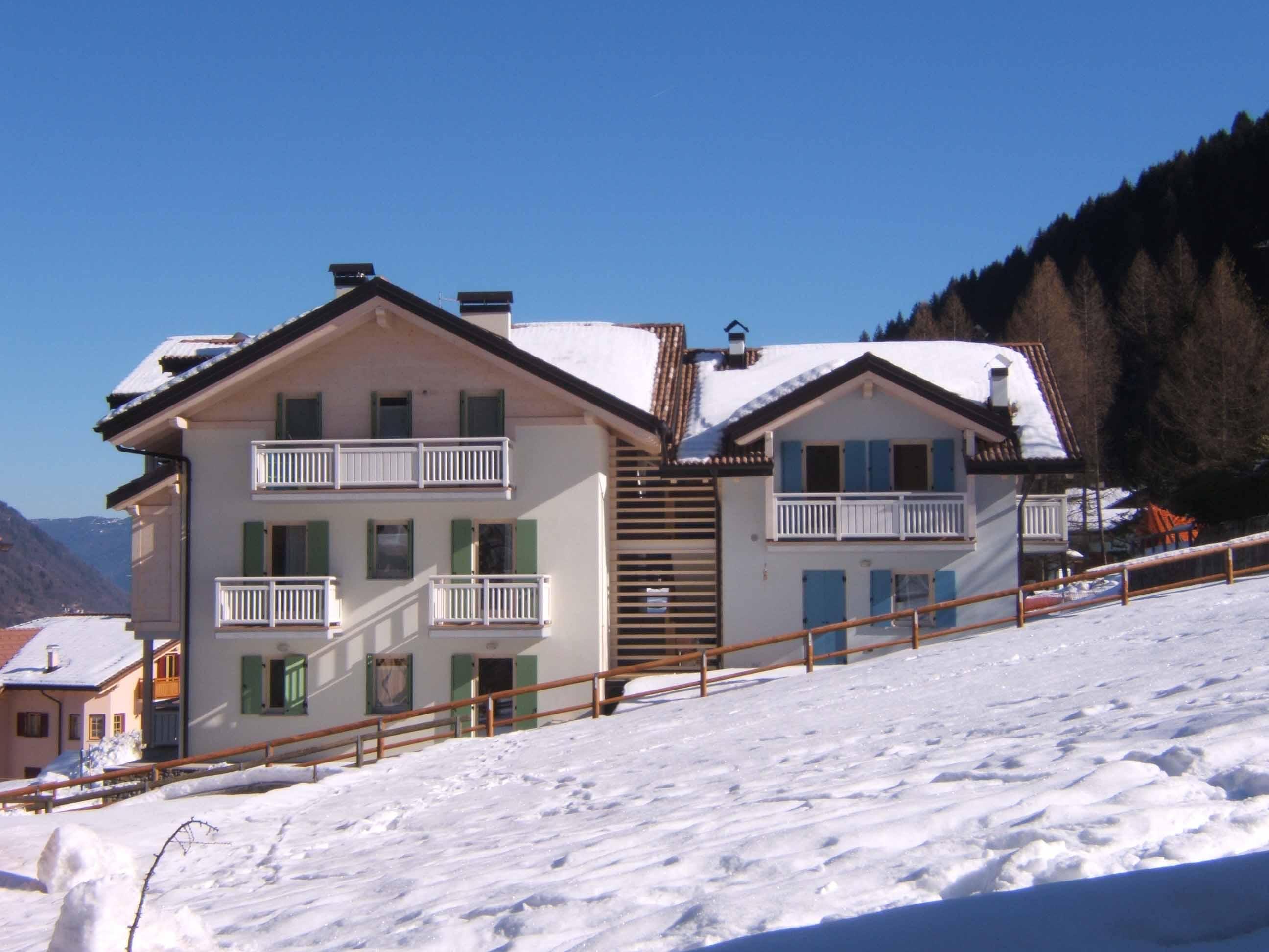 Two wooden and stone buildings covered in snow, surrounded by a mountainous landscape. The sky is clear and blue, creating a tranquil atmosphere.