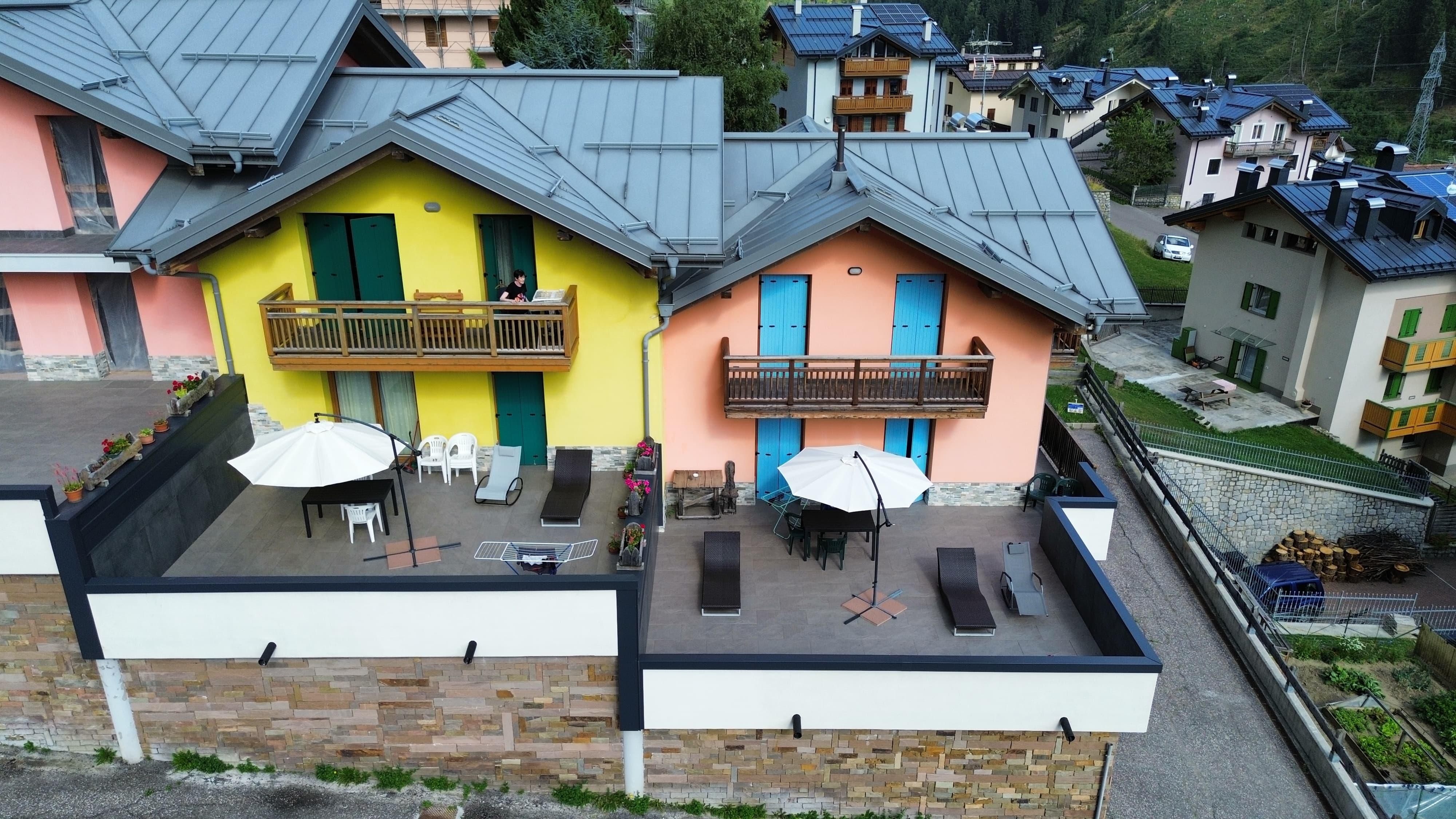 Zwei farbige Häuser mit Balkonen, eines gelb und eines rosa. Der Außenbereich ist mit Tischen und Sonnenschirmen ausgestattet.