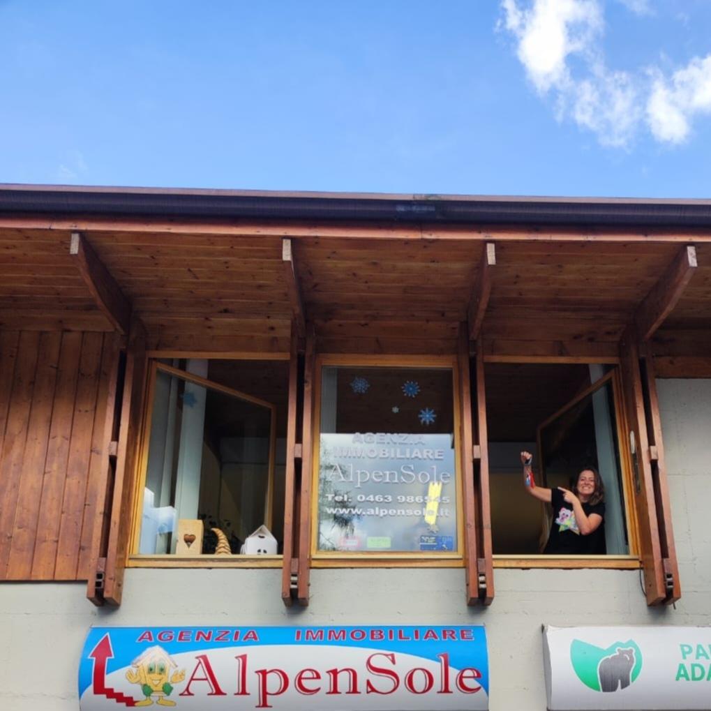 A building with a sign for a real estate agency called AlpenSole. A person is leaning out of an upper floor window, smiling.