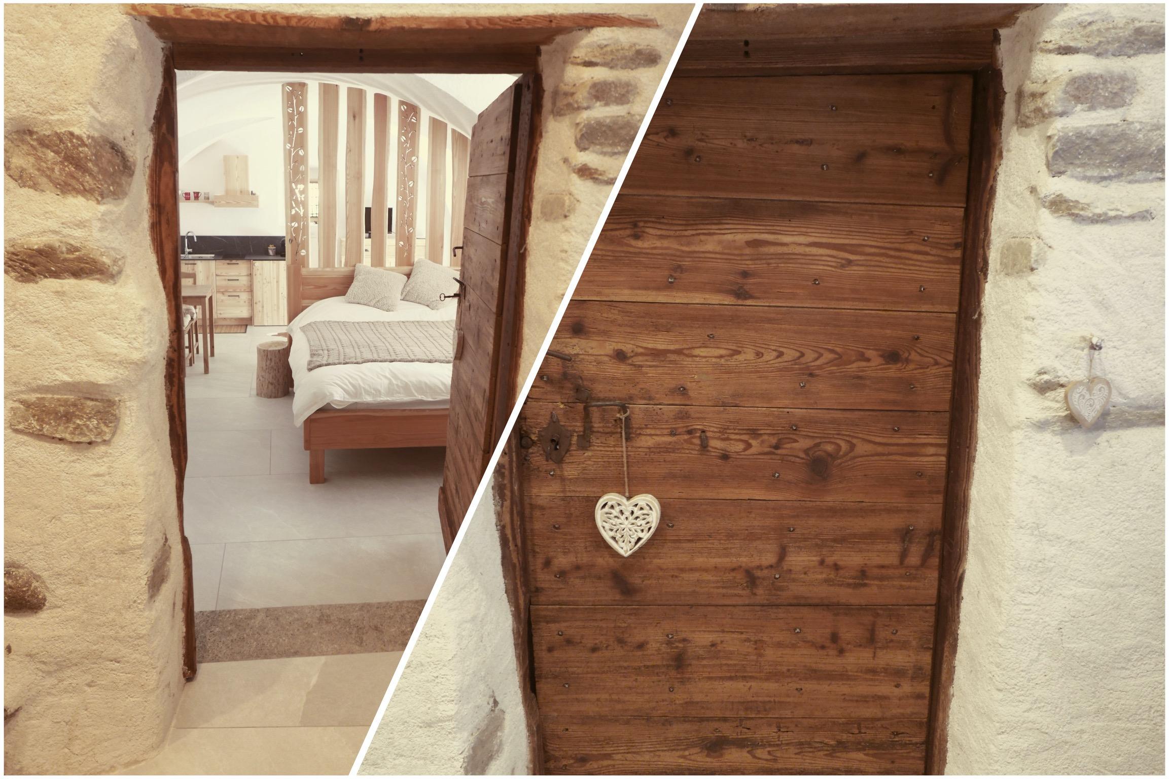 A wooden door with a hanging heart, leading to an elegant bedroom. The room features stone walls and a double bed.