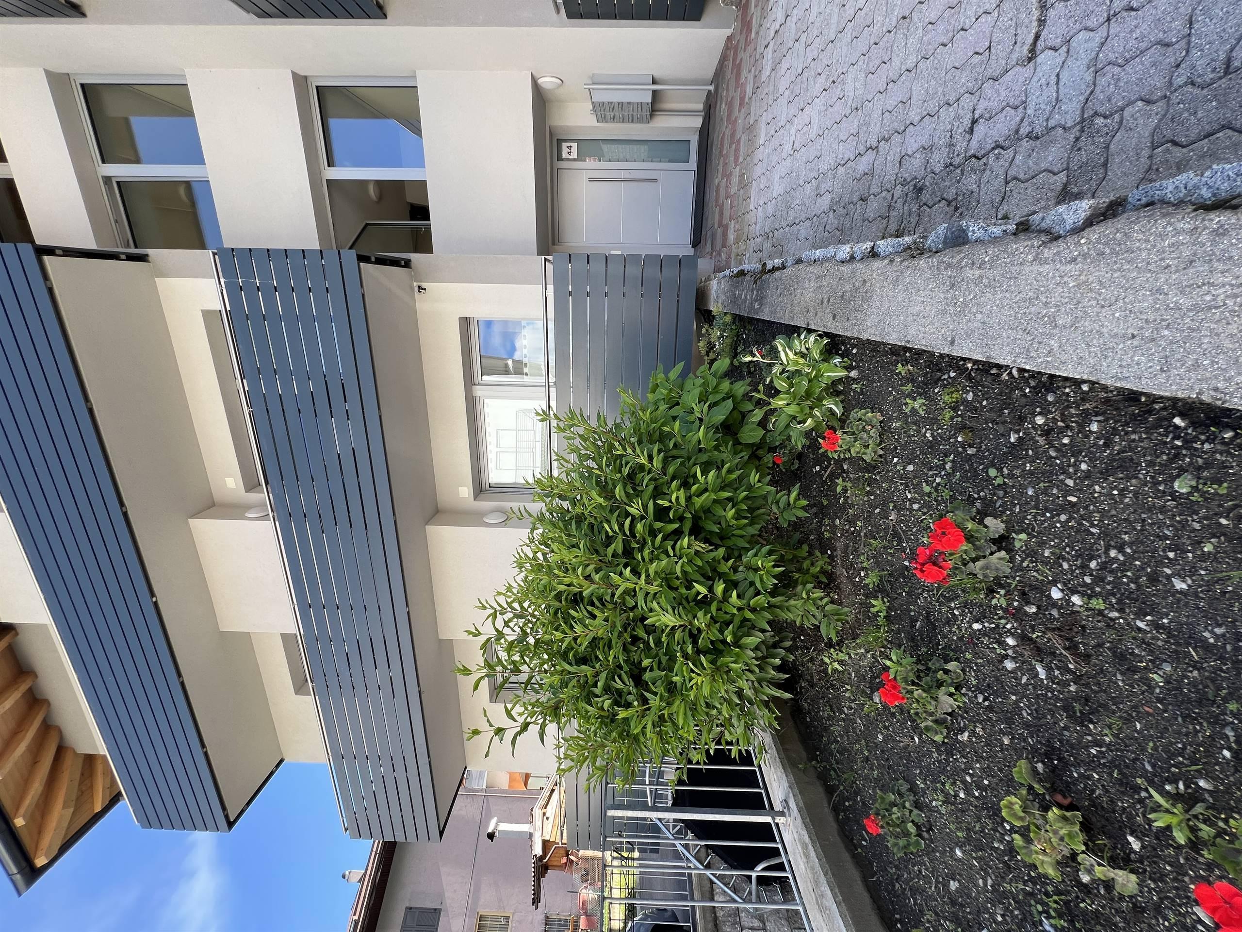 Un edificio moderno con balconi grigi. Davanti ci sono piante verdi e fiori rossi nel giardino.