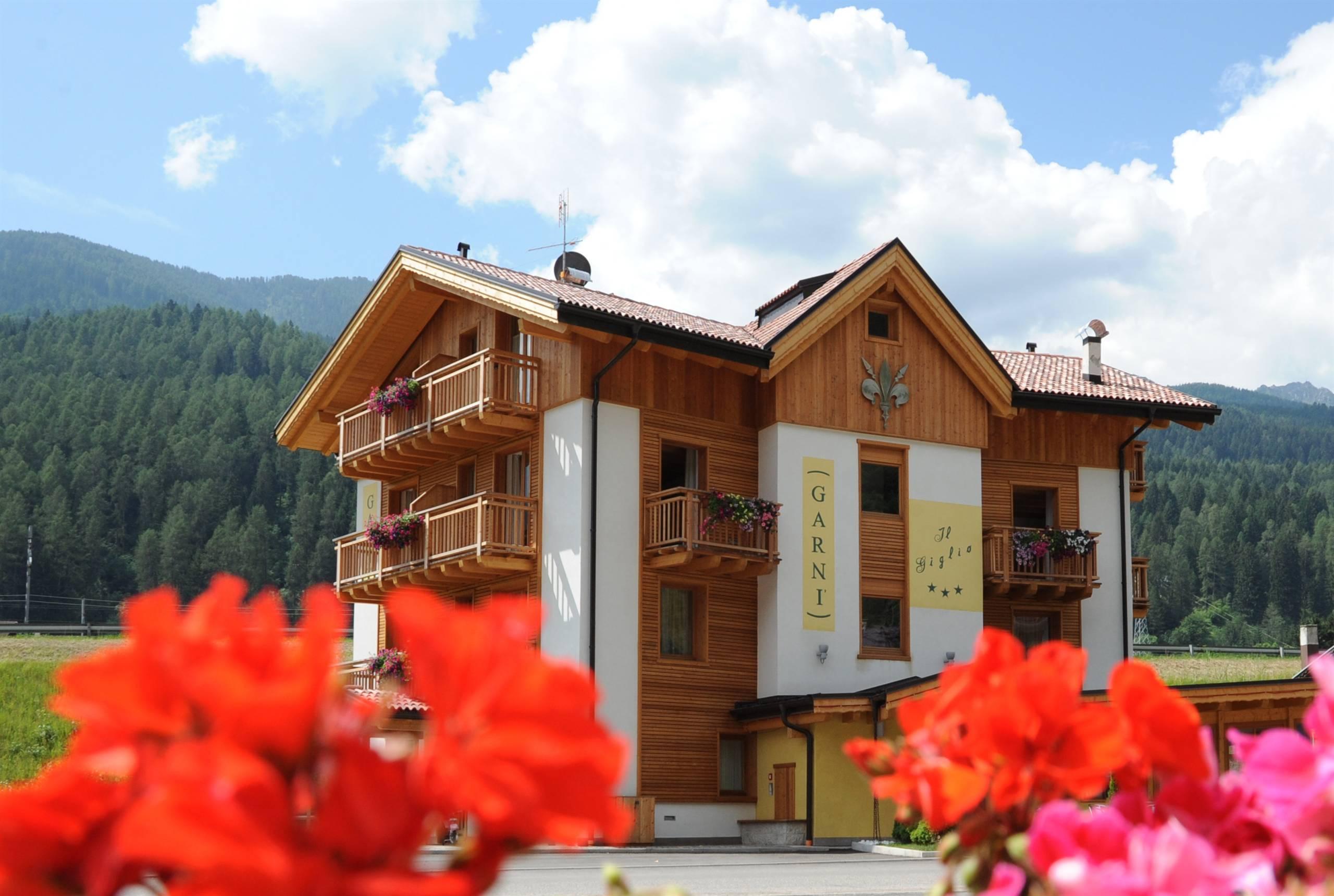 Una casa di legno con balconi e fiori colorati in primo piano. Sullo sfondo si vedono le montagne e un cielo azzurro.