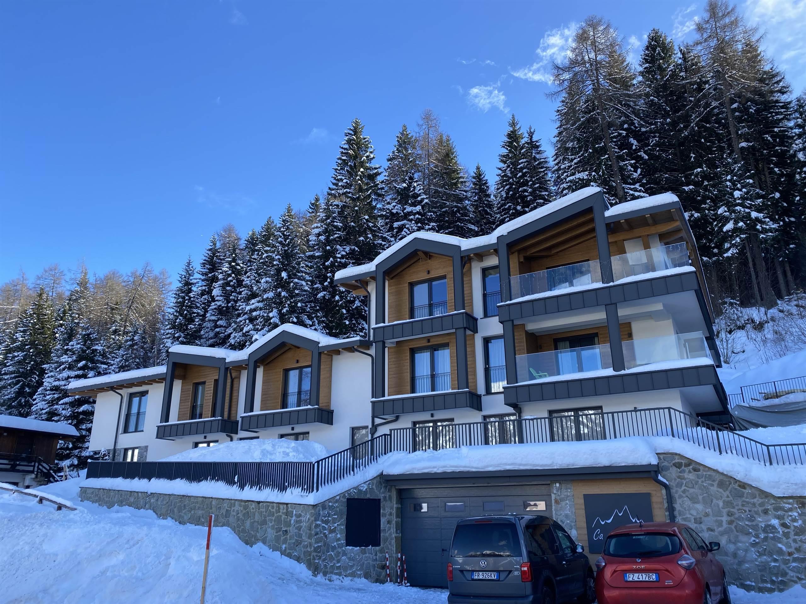 An elegant complex of houses in the snow, surrounded by green trees. The sky is clear and blue, creating a calm and welcoming atmosphere.