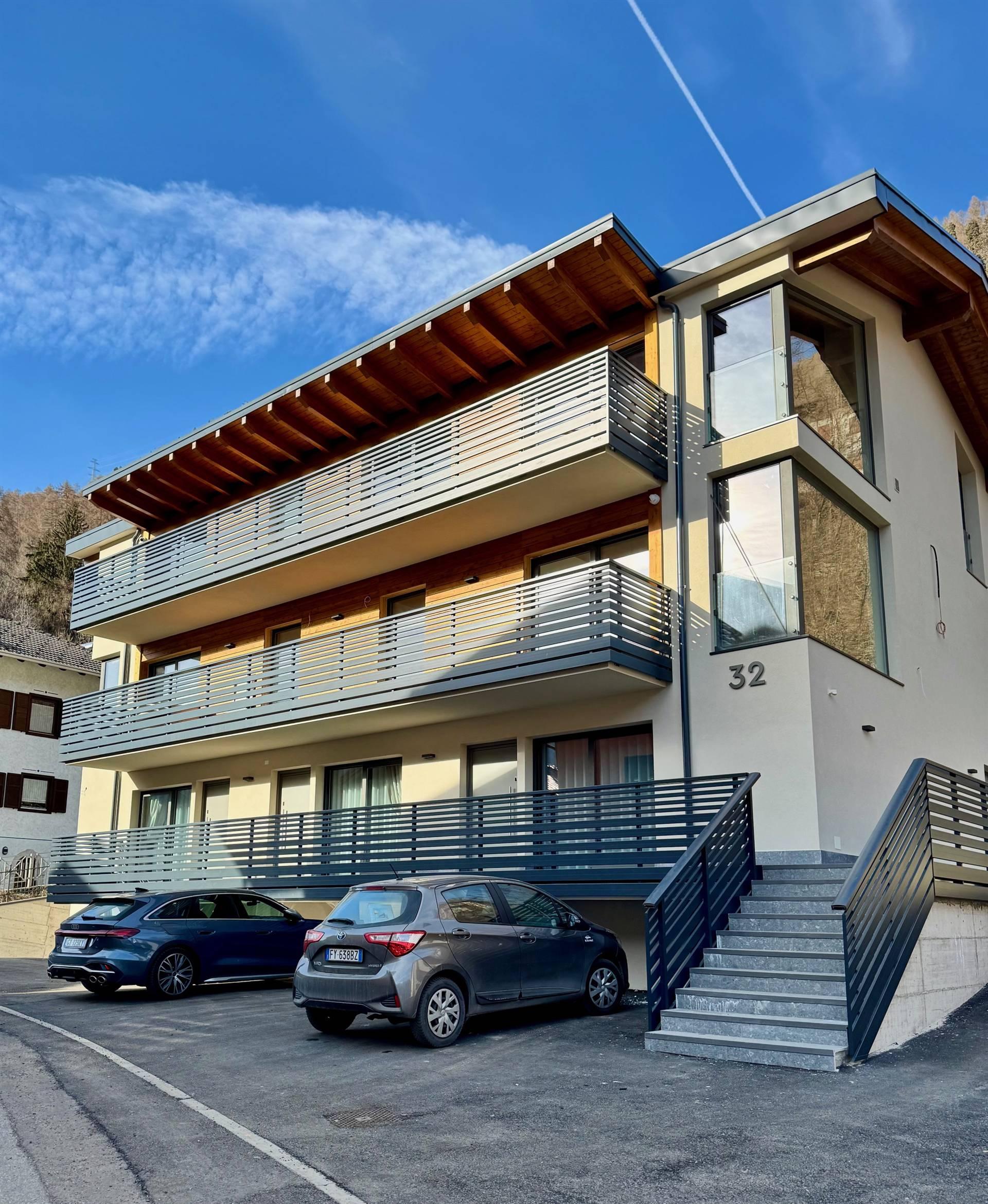 A modern residential building with three floors and balconies. Several cars are parked in front of the entrance.