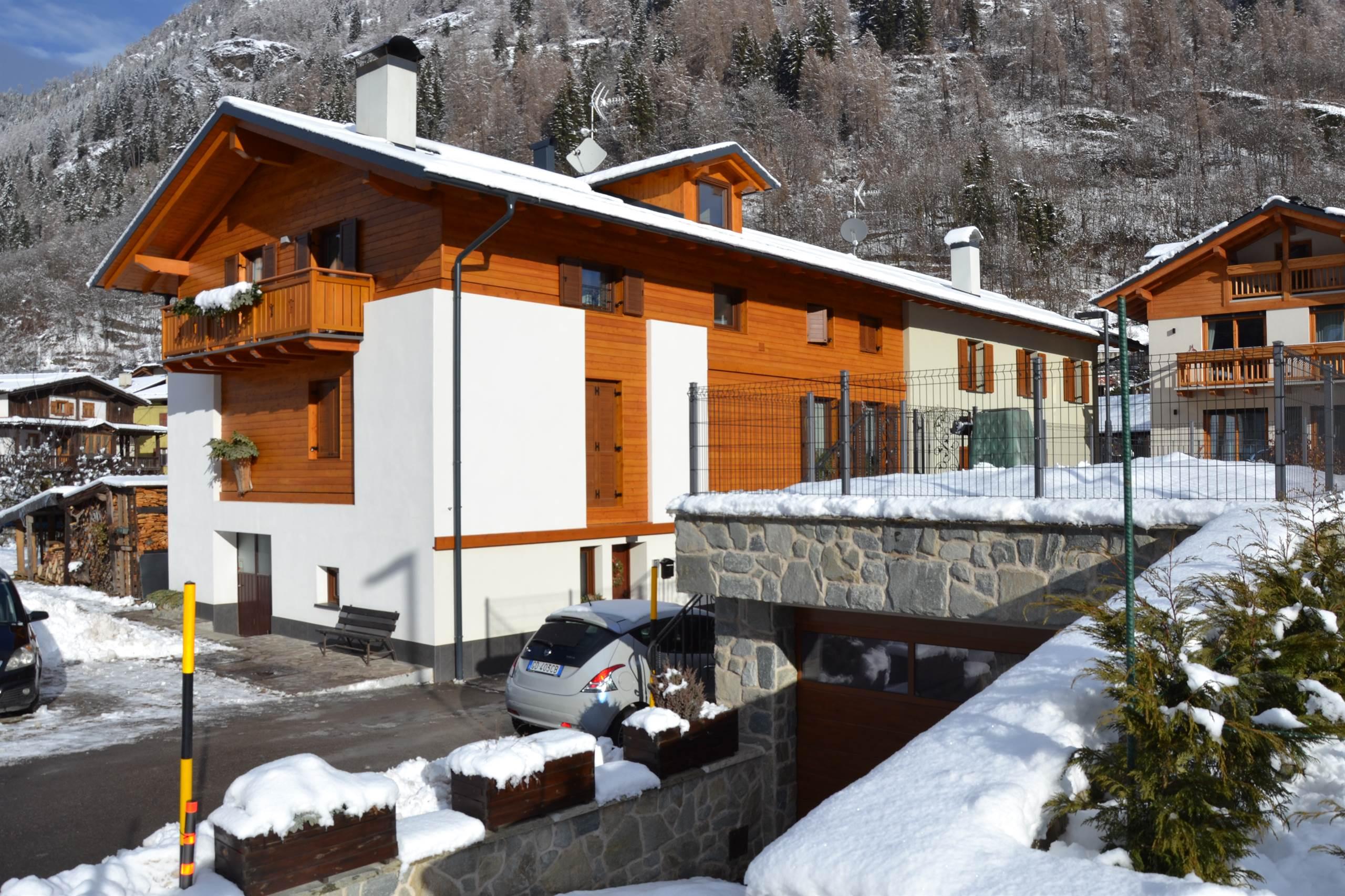 Ein Holzhaus mit Balkonen, umgeben von einer schneebedeckten Landschaft. Im Hintergrund sind weitere Häuser und Berge zu sehen.