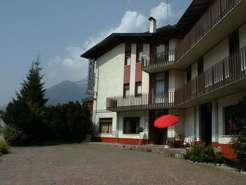 Una vista di un edificio con balconi, circondato da alberi e montagne sullo sfondo. Sotto una delle finestre, c'è un ombrello rosso aperto.