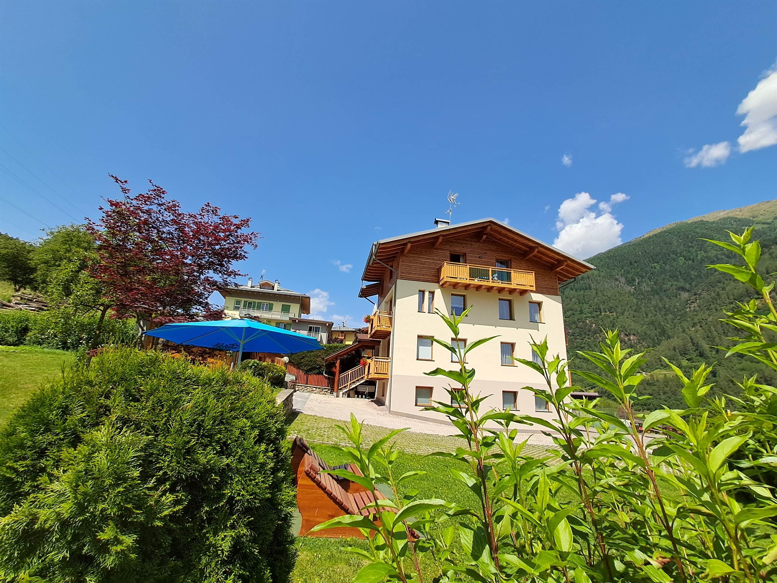 Una casa in montagna circondata da un verde rigoglioso. Sullo sfondo si possono vedere le montagne e un cielo azzurro.