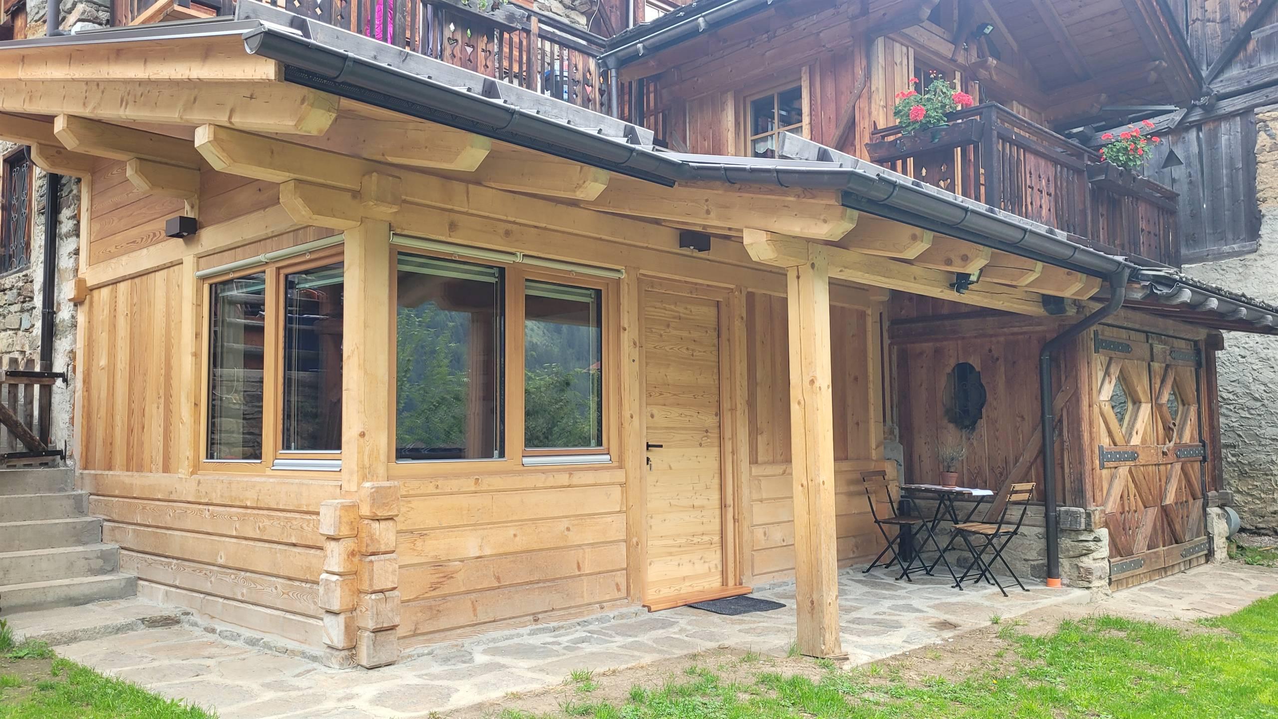 Una casa in legno con finestre ampie, situata in un ambiente montano. Davanti, ci sono due sedie e un tavolino sui ciottoli.