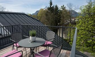 A cozy rooftop terrace with a round table and four chairs. In the background, trees and mountains can be seen under a blue sky.