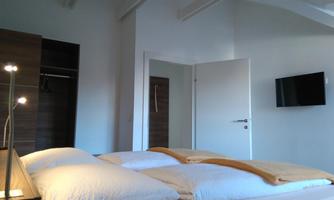 A modern bedroom with a double bed and a TV on the wall. The room has plenty of daylight and a simple, elegant decor.