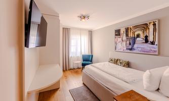 A modern hotel room with a large bed and a flat-screen TV. There is a blue armchair and a window that lets in a lot of natural light.
