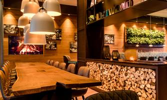 A modern restaurant with a large wooden table and stylish pendant lights. The walls are decorated with plants and photos, creating a cozy atmosphere.