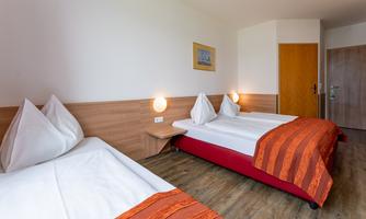 A cozy hotel room with two single beds and modern wooden details. The bedding is white with colorful orange throws.