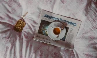 A newspaper lies on a bed with a floral pattern. On it, there is a cup with a small saucer and a spoon.