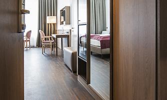 A modern hotel room corridor with wooden flooring and mirrors. In the background, a cozy bedroom with a bed and curtains is visible.
