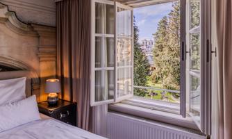 A bedroom with an open window that looks out onto a beautiful garden. The sun shines through the trees, creating an inviting atmosphere.