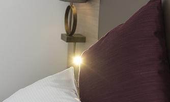A cozy bed with red pillows and a small bedside table. A lamp emits warm light.