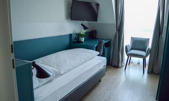 A modern guest room with a single bed, a desk, and a television. The room has large windows that let in plenty of light.