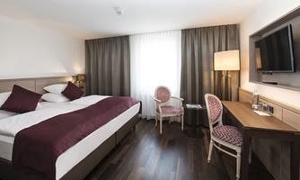 A cozy hotel room with a large bed and elegant decorative elements. The room has a desk, a television, and plenty of natural light.
