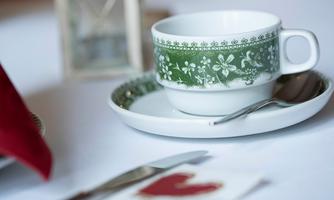 An elegant coffee cup with a floral pattern rests on a saucer. On the table, there is also cutlery and a romantic decoration in the shape of a heart.