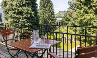 A balcony with a small table and two chairs. In the background, green trees and well-kept gardens are visible.