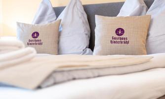 A cozy bed with several pillows and a white blanket. The pillows are printed with the logo of "Gasthaus Hinterbrühl."